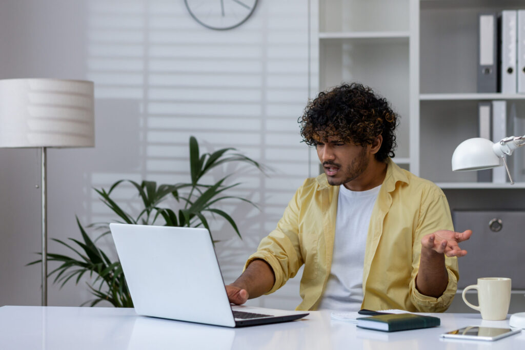 Young man expressing frustration while working on a laptop in a contemporary office environment. The scene captures a stressful moment during a workday.