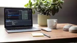 Website maintenance for a small business website - Smart & Simple Communications - Smart & Simple Communications Laptop open on a wooden desk with a code editor on screen, a plant and a coffee mug beside it, the quiet upkeep behind a small business website