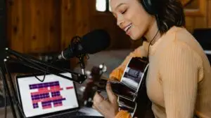what-makes-a-good-jingle - Smart & Simple Communications - Smart & Simple Communications Singer with headphones and an acoustic guitar recording a custom jingle in a warm wood paneled studio, session open on a laptop