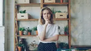 Small business marketing plan: owner thinking through three questions - Smart & Simple Communications - Smart & Simple Communications Small business owner standing in her shop with a hand on her chin, thinking through the three questions in a small business marketing plan